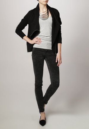 Kvinde iført sort åben jakke, grå ærmeløs top, sorte skinny jeans med syret vask, sorte spidse sko og lagdelt sølvsmykke.