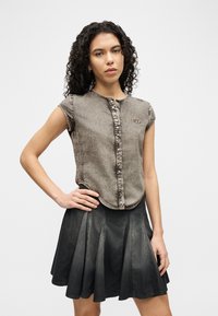 Γυναίκα με κατσαρά μαλλιά που φορά αμάνικο γκρι τοπ με κουμπιά και μαύρη πλεκτή τζιν φούστα, στέκεται με το ένα χέρι στη μέση.