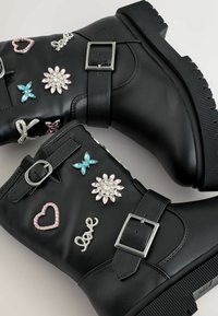 Bocanci negri din piele până la gleznă, cu multiple bijuterii decorative, inclusiv flori, inimi și o flutură, dotati cu catarame din metal și sole texturate.