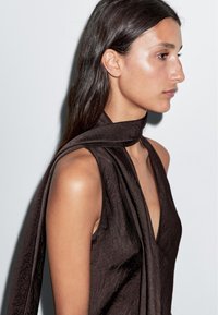 Top senza maniche marrone in tessuto strutturato con scollatura a V profonda e un lungo nodo al collo che pende di lato. Superficie liscia e lucida.