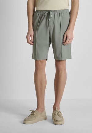 Uomo che indossa pantaloncini verde chiaro con coulisse e scarpe casual beige, in piedi su un pavimento e uno sfondo grigi uniformi.