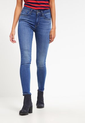 Jean en denim bleu ajusté avec une texture lisse, présentant un style à cinq poches et un léger délavage sur les cuisses, associé à des bottines.