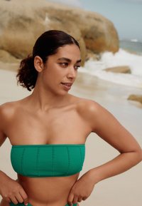 Roheline ribaga bändokujuline bikiinitop dekoratiivse õmblusega, kantud rannal. Liivaline taust ja kaugel paistvad lained.