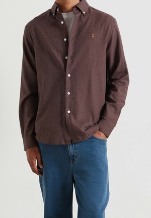 Mann trägt ein braunes Hemd mit Knöpfen über einem weißen T-Shirt und blauen Jeans, eine Hand in der Tasche und die andere locker an der Seite.