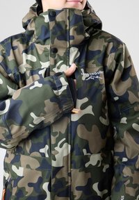 Camouflagejack met schakeringen van groen, bruin en marineblauw; waterdicht materiaal, hoge kraag en voorzakken met ritsen.