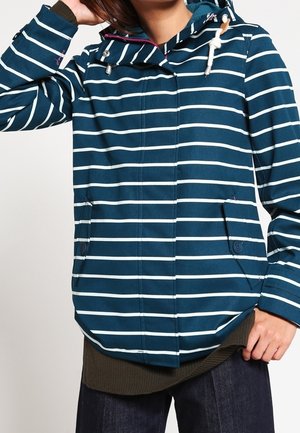 Donna che indossa una giacca con cappuccio a righe orizzontali blu e bianche, con tasche frontali e coulisse, abbinata a pantaloni scuri.