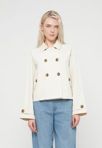 Veste croisée de couleur crème avec de gros boutons marron, poches latérales et larges manches. Tissu lisse avec une coupe courte.