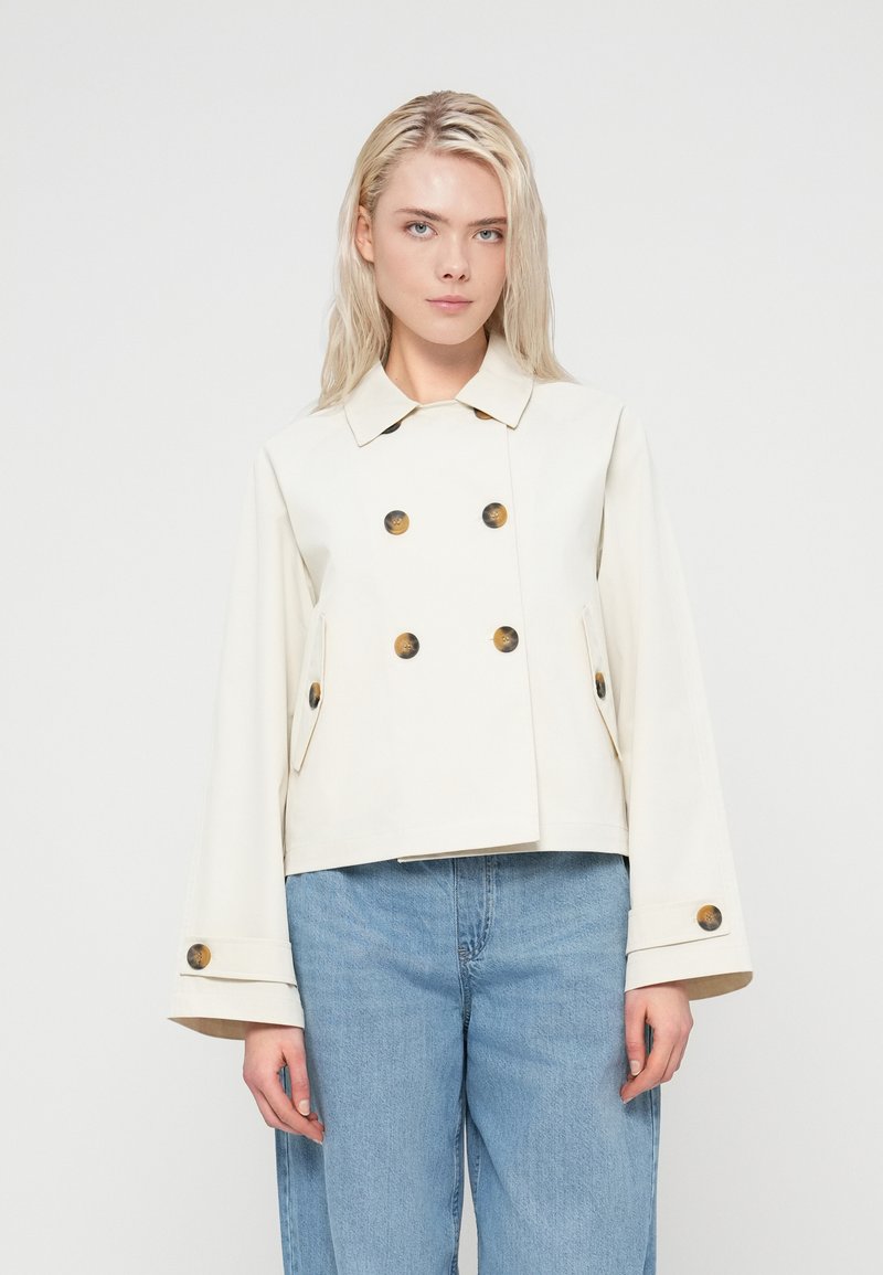 Veste croisée de couleur crème avec de gros boutons marron, poches latérales et larges manches. Tissu lisse avec une coupe courte.