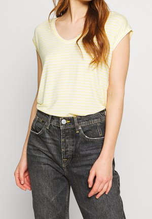 Kobieta w żółto-białej, paskowanej bluzce z krótkim rękawem, włożonej do wysokich spodni jeansowych w kolorze wyblakłej czerni z żółtymi przeszyciami.