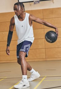 Bela mrežasta košarkaška majica bez rukava, mornarsko modre kratke hlače s bijelim detaljima, crna kompresijska rukava i svijetle sportske cipele. Drži crnu loptu.
