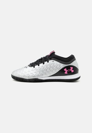 Chaussure de sport blanche et noire à profil bas avec tige texturée, lacets noirs, et logo Under Armour rose sur la zone du talon.