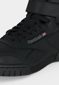 Čierna vysoká teniska Reebok s čiernymi šnúrkami, suchým zipsom a vlajkou Spojeného kráľovstva vedľa loga Reebok na boku.