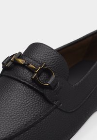 Μαύρο μοκασίνι από δέρμα με υφή, με χάλκινη μεταλλική λεπτομέρεια αλόγου στο επάνω μέρος, ορατή ραφή κατά μήκος των ακμών και σχέδιο χωρίς κορδόνια.