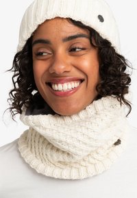 Smilende kvinde med krøllet hår iført en hvid strikbeanie og matchende chunky strikket tørklæde mod en ensfarvet baggrund.