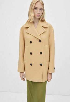 Manteau en mélange de laine beige doux, à double boutonnage avec des boutons noirs, col à revers et poches latérales, associé à une jupe verte.