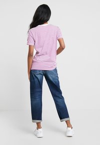 Žena s dlouhými černými vlasy stojí zády, má na sobě růžové a bílé pruhované tričko, zkrácené modré džíny a bílé tenisky.