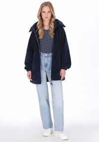 Granatowy płaszcz z futerkiem ekologicznego zapięty na suwak, z kapturem, noszony na paskowanej koszuli. Do kompletu jasnoniebieskie dżinsy i białe trampki.