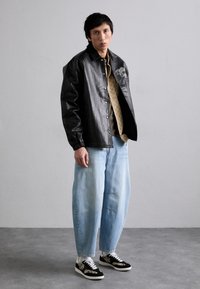 Veste en cuir noire avec boutons-pression, chemise beige à motifs, jean ample bleu clair et baskets noires avec accents floraux.