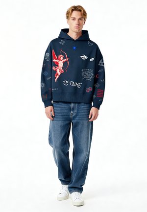 Jeune homme portant un sweat à capuche bleu marine avec un cupidon rouge, des yeux blancs et des graphismes de texte, associé à un jean bleu ample et des baskets blanches, debout devant un fond blanc.