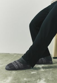 Harmaat ja mustat ruudulliset tossut, joissa pehmeä kangas päällä, tummat kontrastiväri pohjat, ja käytetty harmaiden sukkien kanssa, yhdistettynä mustiin housuihin.