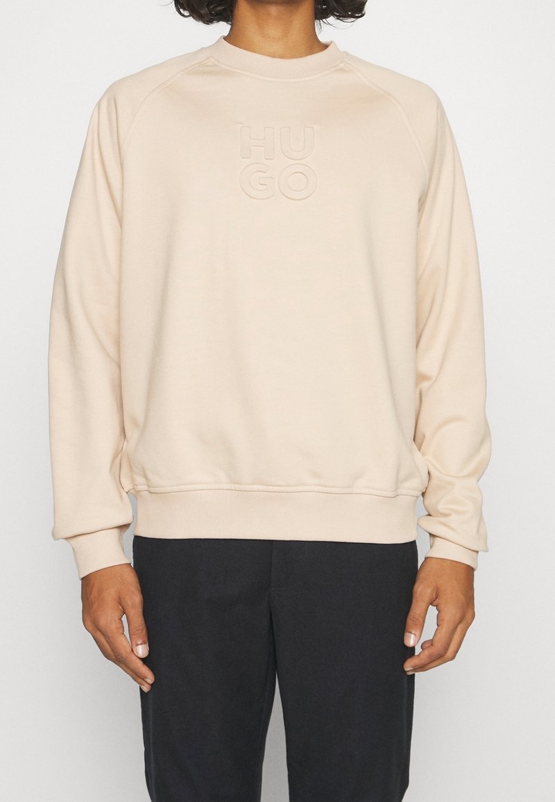 Sudadera de color beige claro con cuello redondo y mangas raglán, que presenta un logo grabado "HUGO" en la parte delantera. Textura suave y corte relajado.