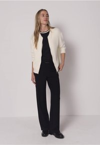 Cardigan blanc texturé porté sur un haut noir et un pantalon noir à jambes larges. Caractérisé par un col arrondi et une coupe décontractée, assorti à des chaussures noires.