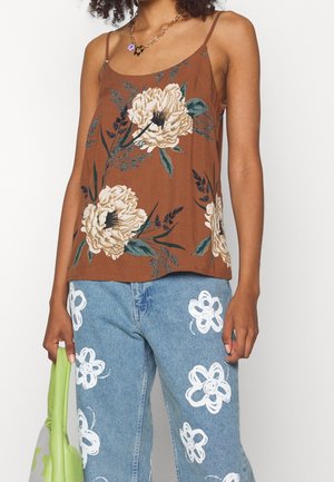 Vrouw draagt een bruine spaghettiband top met bloemenpatroon en een blauwe spijkerbroek met witte bloemenprints, en houdt een lichtgroene handtas vast.