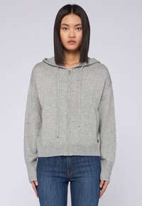 Jasnoszara bluza z zamkiem wykonaną z miękkiej dzianiny, z kapturem na sznurki oraz ściągaczami przy mankietach i u dołu. W połączeniu z niebieskimi dżinsami.