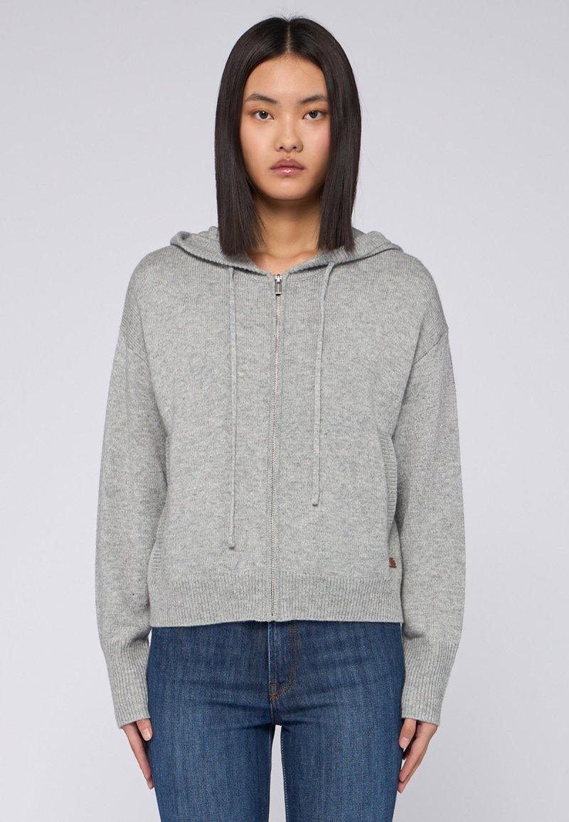 Jasnoszara bluza z zamkiem wykonaną z miękkiej dzianiny, z kapturem na sznurki oraz ściągaczami przy mankietach i u dołu. W połączeniu z niebieskimi dżinsami.