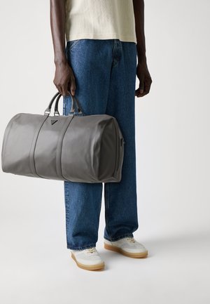 Une personne portant un jean bleu et des baskets blanches tient un grand sac de sport gris avec des poignées doubles sur un fond blanc.