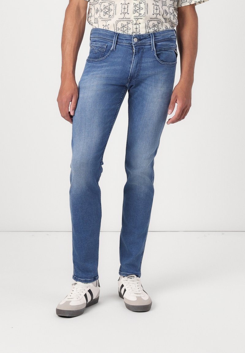Man som har på sig slim-fit blå jeans, mönstrad skjorta instoppad och vita sneakers med svarta randar, stående mot en neutral bakgrund.