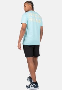 Camiseta azul claro con "WRONG FRIENDS" en texto dorado en la parte posterior, combinada con shorts negros y zapatillas deportivas negras y azules.
