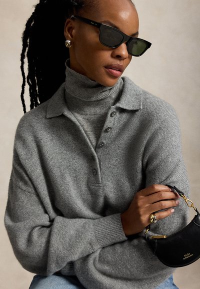 Mujer con gafas de sol negras, suéter gris de cuello alto abotonado, pendientes y anillos de oro, sosteniendo un bolso pequeño negro con detalles dorados.