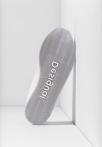 Suela de zapatilla blanca con ondas texturizadas, presentando un prominente logo "Designated" en gris. Acabado suave con elementos de diseño mínimos.