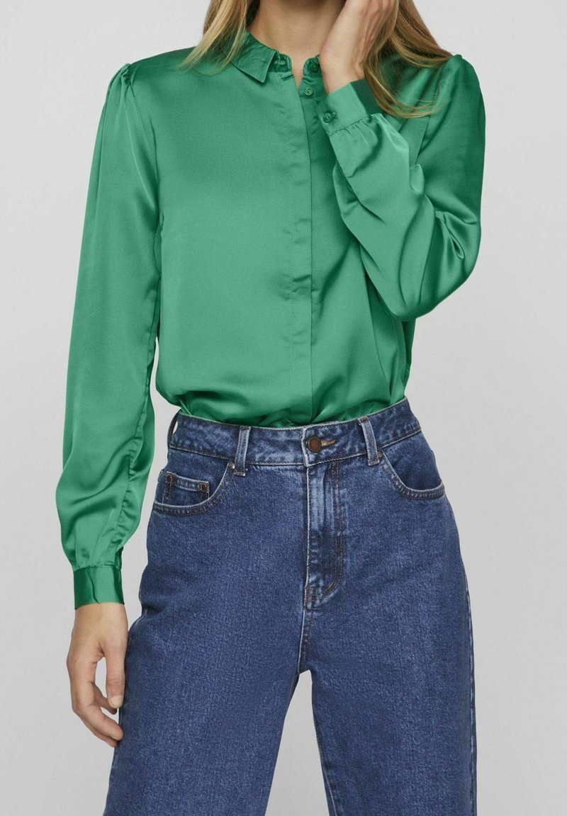 Femme portant un chemisier en satin vert à manches longues, rentré dans un jean en denim bleu taille haute, une main touchant son visage.