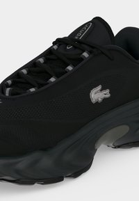Baskets de sport noires en tissu texturé, semelle épaisse, lacets noirs avec accents gris et petit logo crocodile blanc sur le côté.