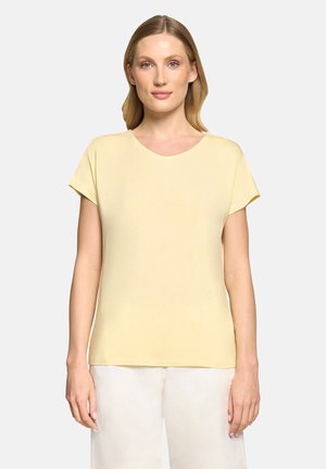 Femme à la peau claire et aux cheveux blonds, portant une chemise jaune pâle à manches courtes et un pantalon de couleur claire, debout devant un fond blanc.