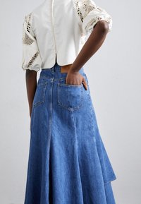 Haut blanc avec des détails en dentelle sur les manches bouffantes, associé à une longue jupe en denim bleu clair avec des poches arrière et un ourlet légèrement évasé.