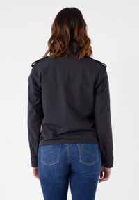 Giacca nera leggera con un design aderente, spalline e bottoni decorativi, abbinata a jeans blu. Texture liscia, dettagli minimi.