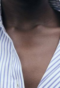 Chemise à rayures bleues et blanches avec col, à encolure en V, partiellement déboutonnée, dévoilant une peau lisse et sombre au niveau du cou et des clavicules.