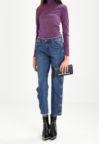 Un col roulé pailleté violet, un jean en denim bleu avec des boutons-pressions sur les côtés, des bottines noires à clous et une pochette noire.