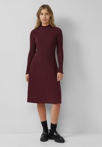 Strickkleid in Burgunderrot mit langen Ärmeln und hohem Halsausschnitt. Figurbetonter Schnitt, der am Saum leicht ausgestellt ist. Getragen mit schwarzen Socken und Loafers.