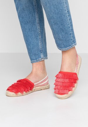 Espadrilles rouges avec un détail de franges en couches et une tige en toile rayée. Les semelles en jute offrent une finition texturée et naturelle. Portées avec un jean bleu.
