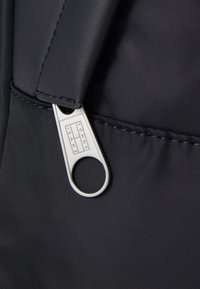 Sac en nylon noir avec tirette argentée arborant un logo "TOMMY JEANS". Texture lisse, coutures renforcées et design épuré.