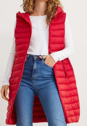 Krullend persoon met een lange rode doorgestikte vest over een wit shirt met lange mouwen en hooggetailleerde blauwe jeans, hand in zak.