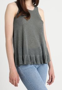 Mörkgrå ärmlös topp i ett glittrande tyg, med en volangkant och en avslappnad passform. Bärs med ljusblå jeans.