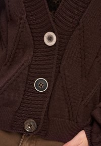 Brązowy sweter z dzianiny z prążkowaną teksturą, dużymi skośnymi wzorami warkoczowymi oraz trzema guzikami: przezroczystym, czarnym i brązowym marmurkowym.