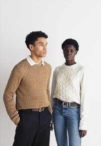 To personer står ved siden av hverandre. Den ene har på seg en teksturert brun genser og svarte bukser; den andre har på seg en kremfarget genser med flettemønster og jeans.
