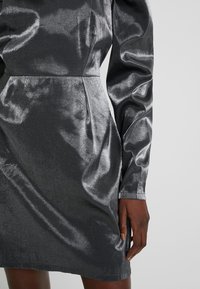 Robe longue à manches longues en argent métallique brillant avec manches légèrement bouffantes et taille cintrée, portée par une personne à la peau foncée.