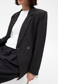 Melna pielāgota žakete ar plecu spilventiņiem, ar vienu pogu un teksturētu audumu. Valkāta virs baltas blūzes un kombinēta ar melnām biksēm.